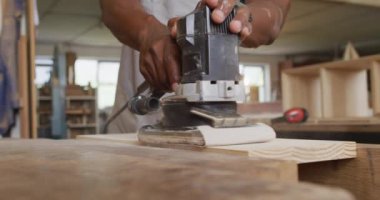 Mid section of african american male carpenter using an electric grinder to grind wooden plank. carpentry, craftsmanship and handwork concept