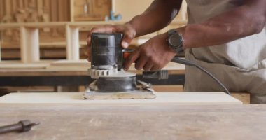 Mid section of african american male carpenter using an electric grinder to grind wooden plank. carpentry, craftsmanship and handwork concept