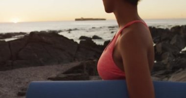 Gün batımında deniz kenarındaki kayalıklarda yoga minderi tutan Afrikalı Amerikalı kadın portresi. sağlıklı yaşam tarzı ve yaşam konsepti