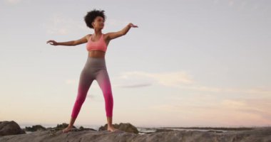 Afrika kökenli Amerikalı kadın gün batımında deniz kenarındaki kayalarda yoga ve meditasyon yapıyor. sağlıklı yaşam tarzı ve yaşam konsepti