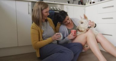 Kafkasyalı lezbiyen çift ellerinde kahve fincanlarıyla evde mutfakta köpekleriyle oynuyorlar. lgbt ilişkisi ve yaşam tarzı konsepti