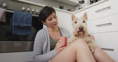 Kafkasyalı kadın elinde kahve fincanıyla evde mutfakta köpeğiyle oynuyor. evcil hayvan sevgisi ve bakım kavramı