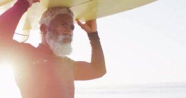 Kıdemli Afro-Amerikan adam güneşli sahilde sörf tahtasıyla yürüyor. sağlıklı ve aktif zaman plaj tatili.