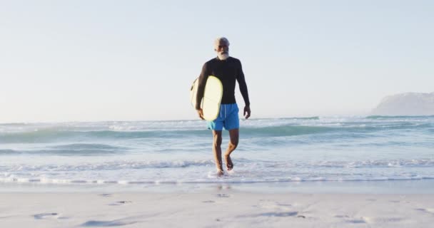 上了年纪的非洲裔美国人和冲浪板一起在阳光灿烂的海滩上散步 健康而活跃的海滨假期 — 图库视频影像