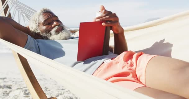 快乐的资深非洲裔美国人读书 躺在阳光灿烂的海滩上的吊床上 健康而活跃的海滨假期 — 图库视频影像
