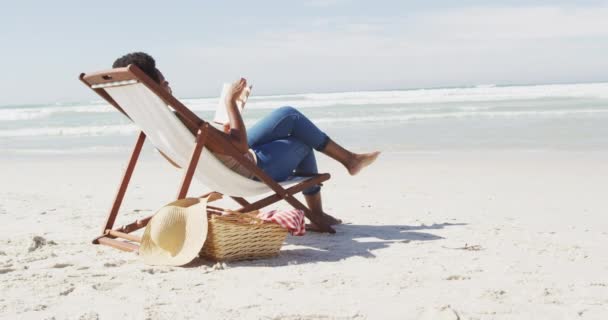 非洲裔美国妇女在阳光灿烂的海滩上躺在日光浴床上看书 健康而活跃的海滨假期 — 图库视频影像