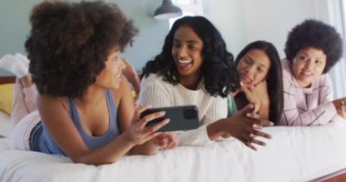 Yatakta uzanan ve yatak odasında akıllı telefon kullanan mutlu bayan arkadaşlar. evde kaliteli zaman geçirmek.