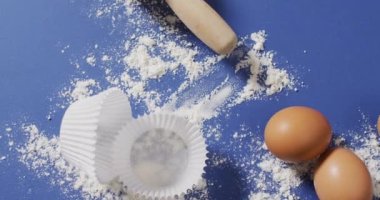 Pişirme malzemeleri, kek kağıtları, yumurtalar ve aletlerin mavi yüzeyde un ile yattığı video. Pişirme, yemek hazırlama, tat ve lezzet kavramı.