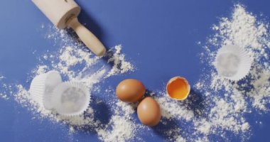 Pişirme malzemeleri, kek kağıtları, yumurtalar ve aletlerin mavi yüzeyde un ile yattığı video. Pişirme, yemek hazırlama, tat ve lezzet kavramı.