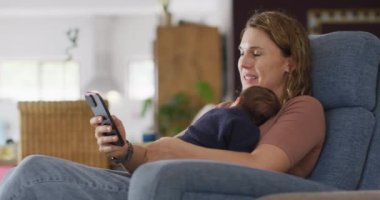 Kafkasyalı annenin yeni doğmuş bir bebekle koltukta oturup akıllı telefon kullandığı bir video. annelik, ebeveyn sevgisi ve yeni doğan bebek konseptiyle ilgilenmek dijital olarak üretilmiş video.