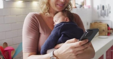 Kafkasyalı anne ve yeni doğmuş bebeği bebek arabasında mutfaktaki akıllı telefonu kullanırken çekilmiş bir video. annelik, ebeveyn sevgisi ve yeni doğan bebek konseptiyle ilgilenmek dijital olarak üretilmiş video.