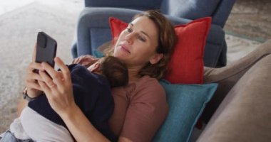 Akıllı telefonlu beyaz bir annenin yeni doğmuş bir bebekle kanepede yattığı bir video. annelik, ebeveyn sevgisi ve yeni doğan bebek konseptiyle ilgilenmek dijital olarak üretilmiş video.