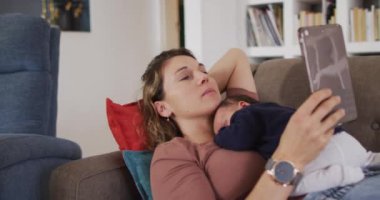 Kafkasyalı bir annenin tabletle kanepeye uzanmış yeni doğmuş bir bebekle birlikte yattığı video. annelik, ebeveyn sevgisi ve yeni doğan bebek konseptiyle ilgilenmek dijital olarak üretilmiş video.