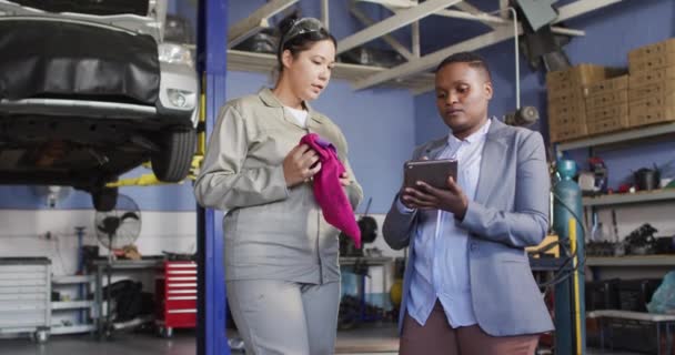 自動車部品を検索するタブレット付きの2つの多様な女性の車の整備士やビジネスオーナーのビデオ 車修理工場で働き女性らしい小さなビジネスコンセプトを運営しています — ストック動画