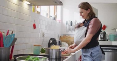 Kafkasyalı anne ve yeni doğmuş bebeği bebek taşıyıcısında mutfakta yemek pişirirken çekilmiş bir video. annelik, ebeveyn sevgisi ve yeni doğan bebek konseptiyle ilgilenmek dijital olarak üretilmiş video.