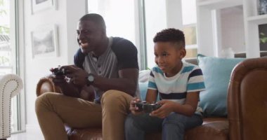 Video of happy african american father and son sitting on sofa and playing video games. Family, spending quality time together at home with technology concept.