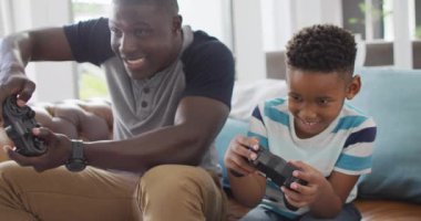 Video of happy african american father and son sitting on sofa and playing video games. Family, spending quality time together at home with technology concept.