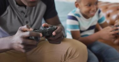 Video of happy african american father and son sitting on sofa and playing video games. Family, spending quality time together at home with technology concept.