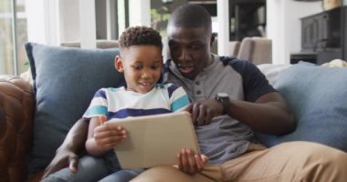 Video of happy african american father and son sitting on sofa and using tablet. Family, spending quality time together at home with technology concept.
