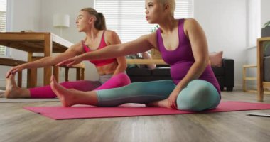 Happy diverse female couple doing yoga together in living room, stretching. Domestic lifestyle, spending free time at home.