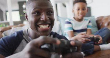 Video of happy african american father and son sitting on sofa and playing video games. Family, spending quality time together at home with technology concept.