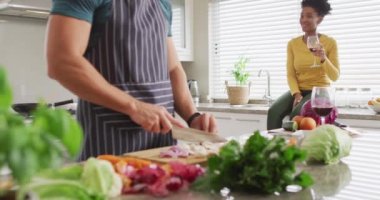Video of happy diverse couple preparing meal, drinking wine and having fun in kitchen. Love, relationship and spending quality time together at home.