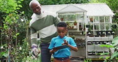 Torunuyla birlikte bitkilere bakan, bahçede tablet kullanan mutlu kıdemli Afrikalı Amerikalı adam. Dışarıda vakit geçirmek, bahçede çalışmak..