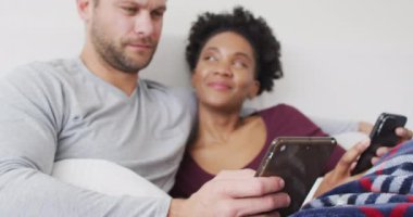 Video of happy diverse couple using smartphone in bed and talking. Love, relationship and spending quality time together at home.