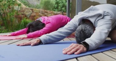 Happy diverse couple doing yoga, stretching on yoga mats at terrace. Spending quality time at home.