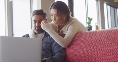 Happy diverse couple sitting in living room using laptop and talking. Spending quality time at home.