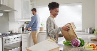 Video of happy diverse couple unpacking groceries in kitchen. Love, relationship and spending quality time together at home.