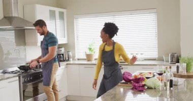 Video of happy diverse couple dancing in kitchen in aprons. Love, relationship and spending quality time together at home.