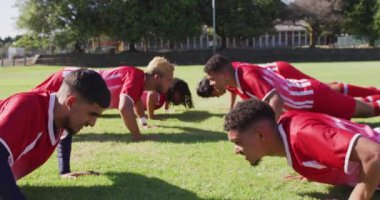 Diverse group of male football players warming up on field, doing push-ups. Male football team, inclusivity and fitness in team sports.