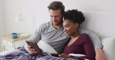 Video of happy diverse couple using tablet in bed and talking. Love, relationship and spending quality time together at home.