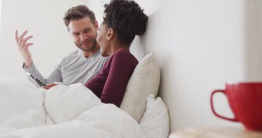 Video of happy diverse couple using tablet in bed and talking. Love, relationship and spending quality time together at home.