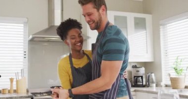 Video of happy diverse couple dancing in kitchen in aprons. Love, relationship and spending quality time together at home.