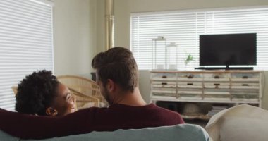 Video of back view of happy diverse couple drinking coffee and watching tv with copy space. Love, relationship and spending quality time together at home.