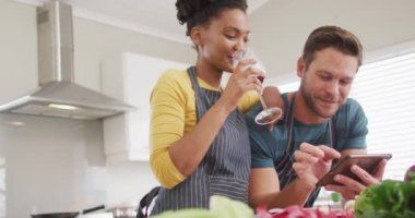 Video of happy diverse couple preparing meal, using tablet and drinking wine in kitchen. Love, relationship and spending quality time together at home.