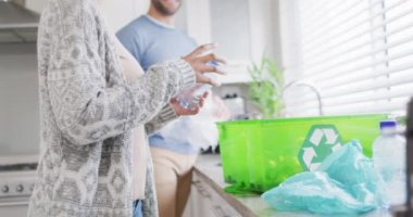 Video of happy diverse couple collecting plastic bottles for recycling at home. Relationship, living together, recycling and eco awareness concept digitally generated video.