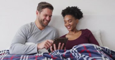 Video of happy diverse couple using tablet in bed and talking. Love, relationship and spending quality time together at home.