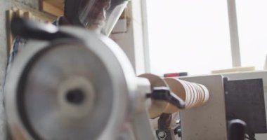 African american male carpenter wearing protective helmet turning wood on a lathe at carpentry shop. Carpentry, craftsmanship and handwork concept