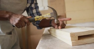 Mid section of african american male carpenter hammering nails into the wood at a carpentry shop. Carpentry, craftsmanship and handwork concept