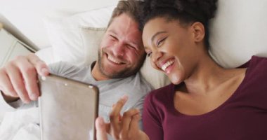 Video of happy diverse couple using tablet in bed and talking. Love, relationship and spending quality time together at home.