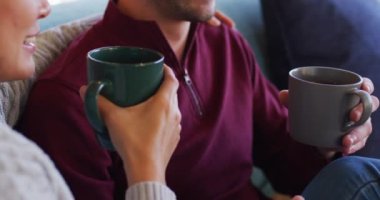 Video of back view of happy diverse couple sitting on sofa and drinking coffee. Love, relationship and spending quality time together concept.