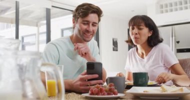 Video of happy diverse couple using smartphone and drinking coffee together in kitchen. Love, relationship and spending quality time together concept.
