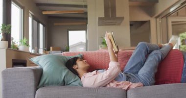 Video of biracial woman lying on sofa and reading book. Relax, leisure and spending free time at home with technology concept.
