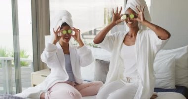 Video of happy diverse female friends in robes having fun with cucumber slices. Friendship, taking care of yourself and beauty concept.