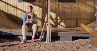 Video of happy caucasian female skateboarder resting and using smartphone. Skateboarding, sport, active lifestyle and hobby concept.