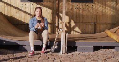 Video of happy caucasian female skateboarder resting and using smartphone. Skateboarding, sport, active lifestyle and hobby concept.