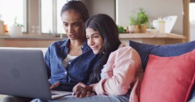 Video of happy diverse female friends using laptop on sofa. Friendship, spending quality time together at home.